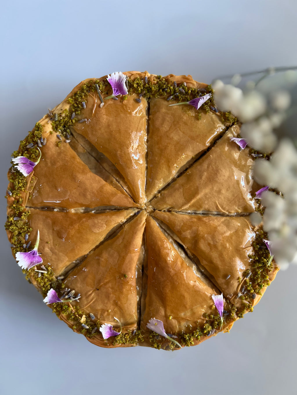 A top-down view of a sliced round pastry, likely baklava or a similar layered cake, with glossy brown filling, green pistachio-like crust around the edge, and small purple flower petals as garnish, set against a pale blue background.