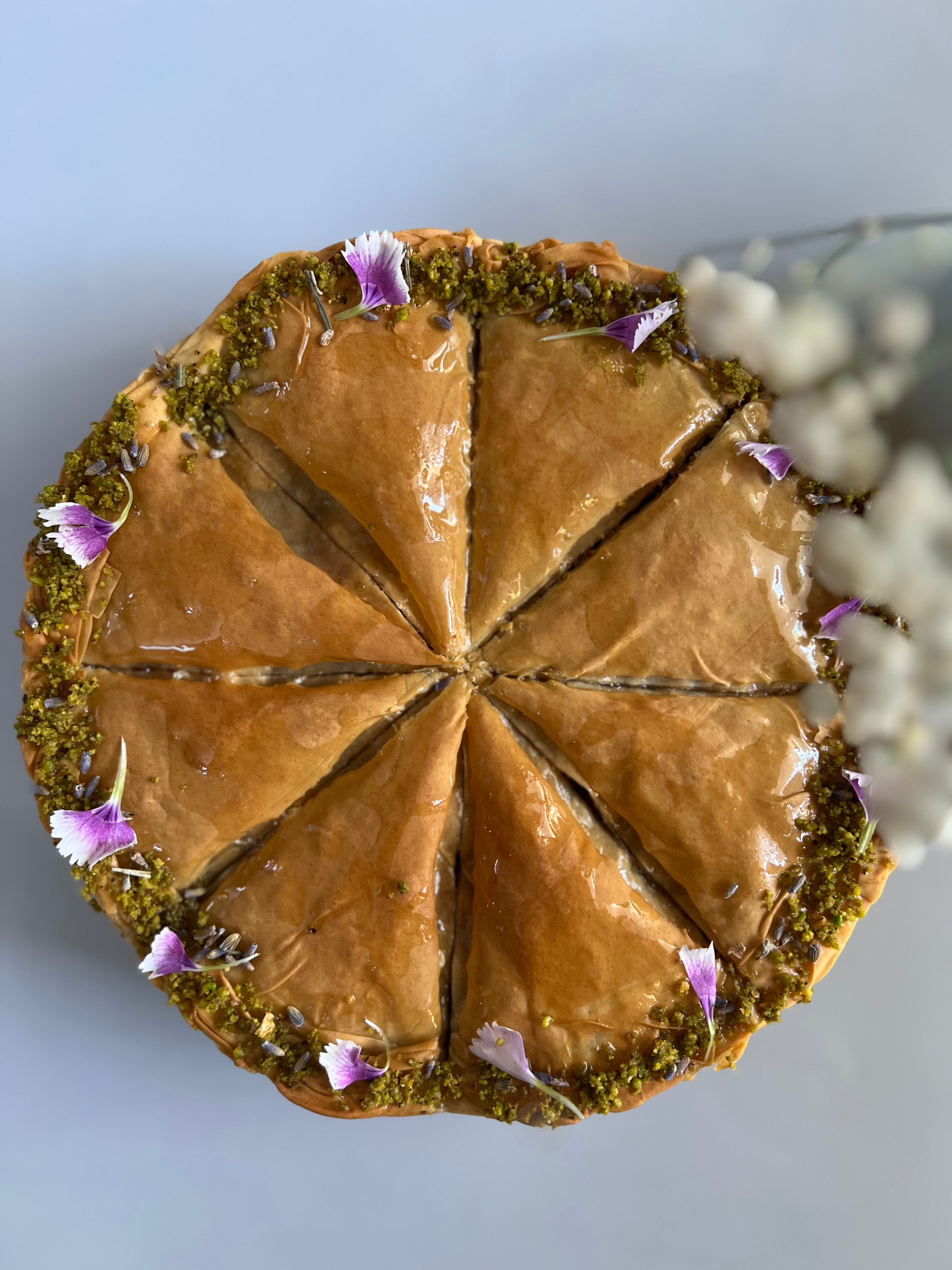 A top-down view of a sliced round pastry, likely baklava or a similar layered cake, with glossy brown filling, green pistachio-like crust around the edge, and small purple flower petals as garnish, set against a pale blue background.