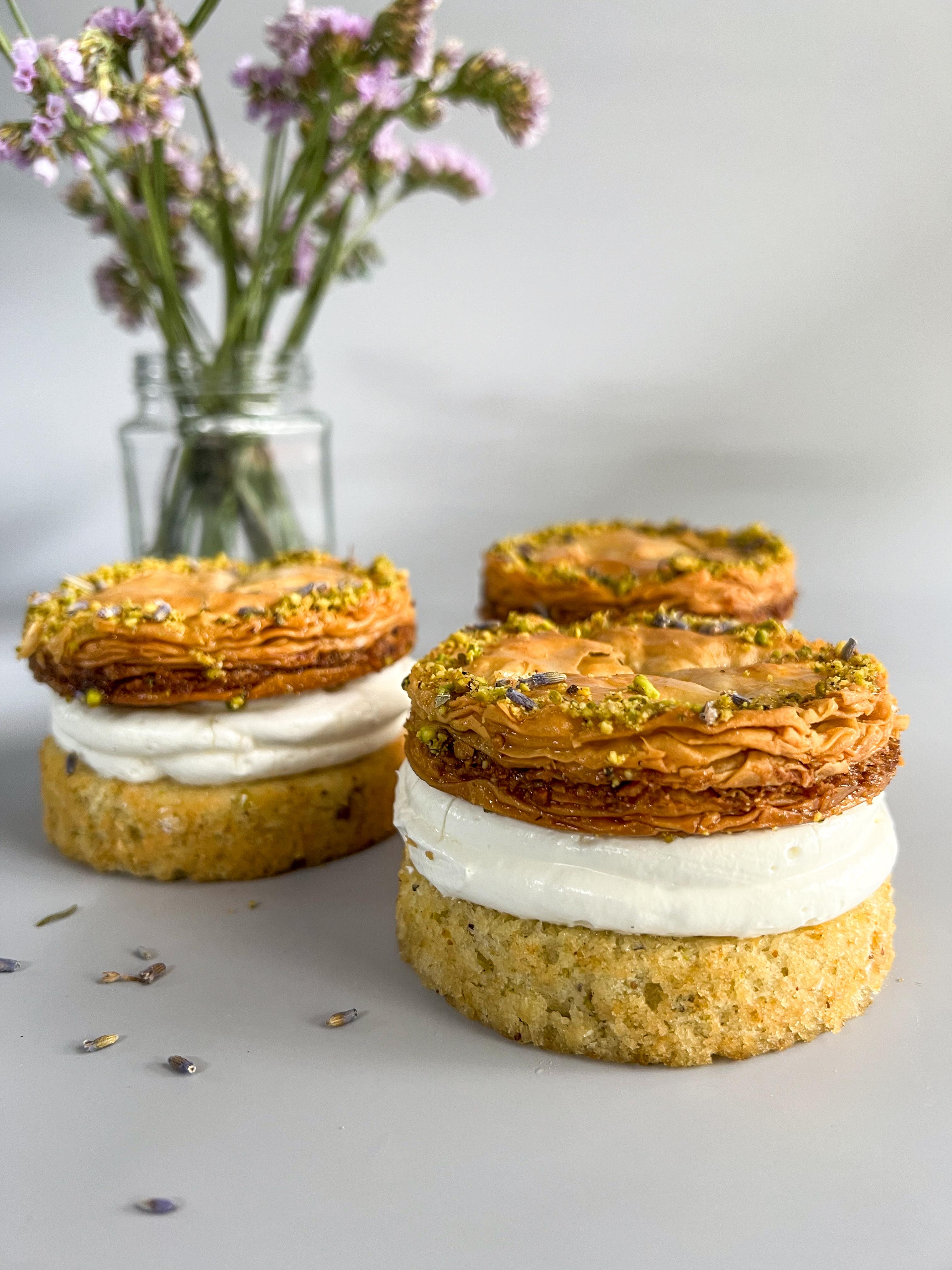 Three layered pastries with golden-brown pastry, white cream, and pistachio topping, arranged on a light gray surface with a small glass vase of flowers in the background.