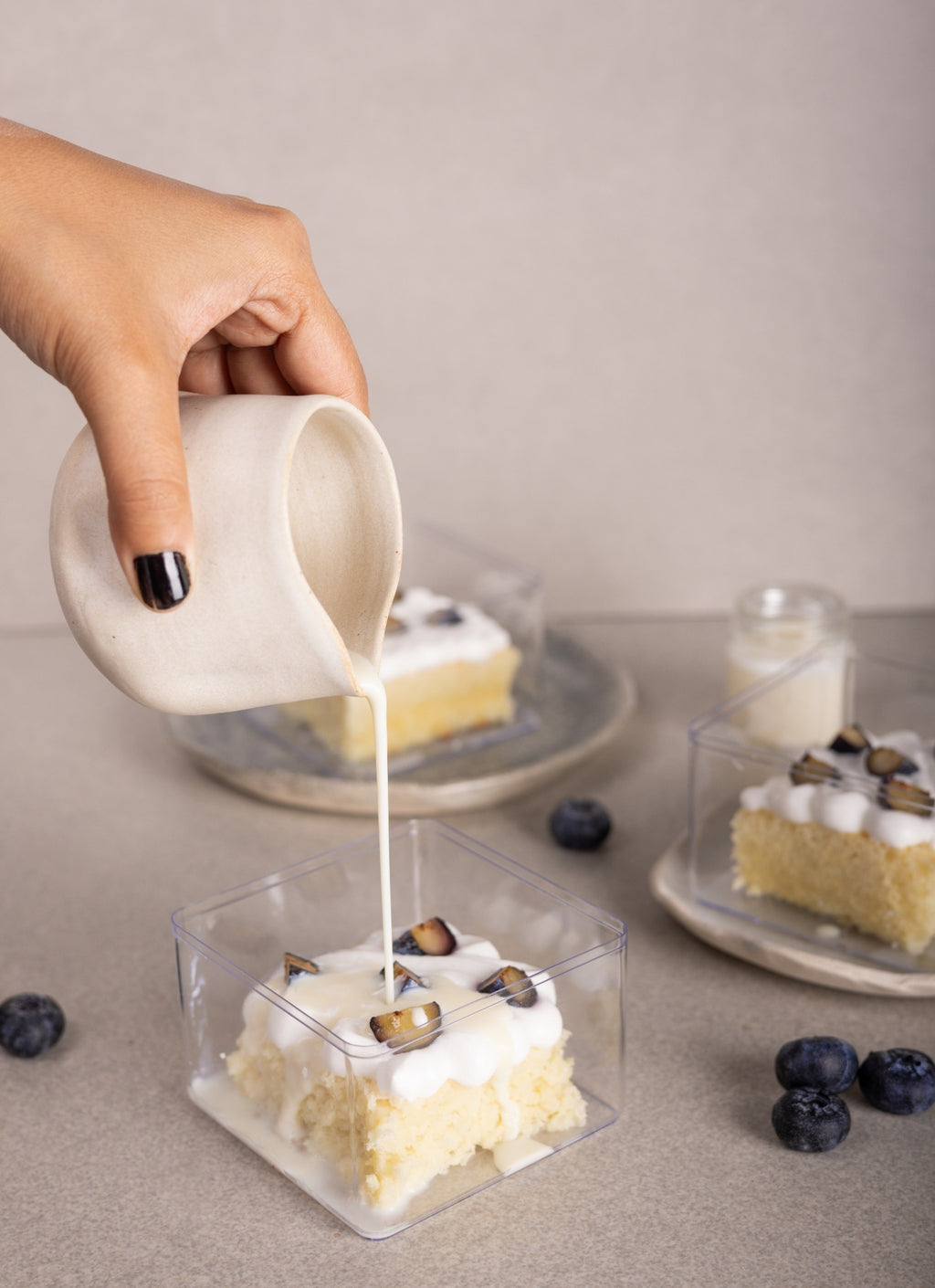 A hand with black nail polish pours a creamy white sauce from a small white ceramic pitcher onto a square clear plastic container holding a slice of Tres Lechese topped with white frosting and bluberry pieces. Two more similar dessert containers sit on plates in the softly blurred background on a neutral beige surface.