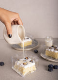 A hand with black nail polish pours a creamy white sauce from a small white ceramic pitcher onto a square clear plastic container holding a slice of Tres Lechese topped with white frosting and bluberry pieces. Two more similar dessert containers sit on plates in the softly blurred background on a neutral beige surface.