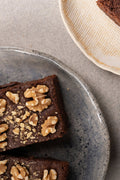 Chocolate Walnut Brownie topped with sliced walnuts, served on a speckled gray ceramic plate on a neutral gray surface, with a partial view of another plate in the top right corner.