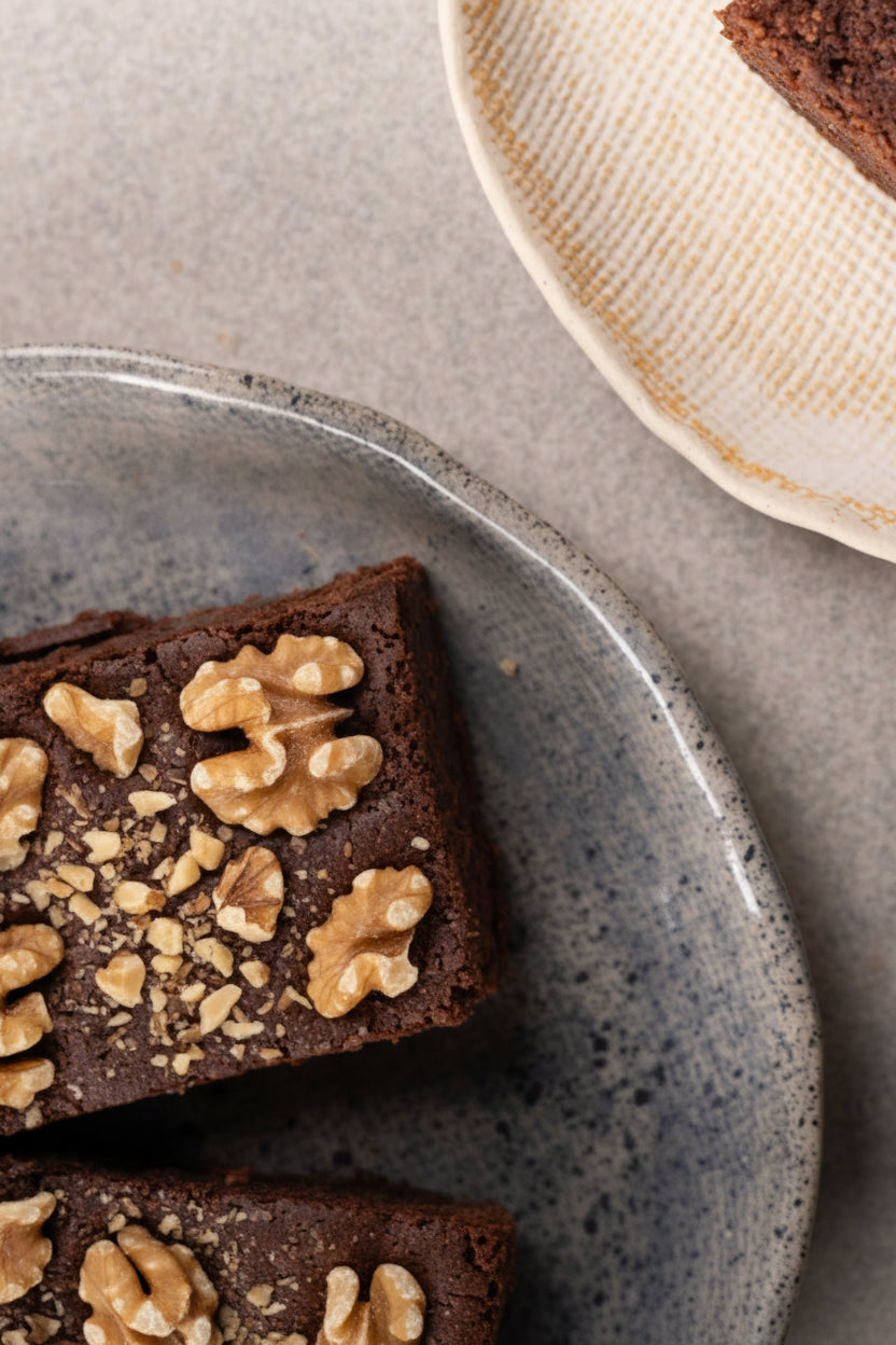 Chocolate Walnut Brownie topped with sliced walnuts, served on a speckled gray ceramic plate on a neutral gray surface, with a partial view of another plate in the top right corner.