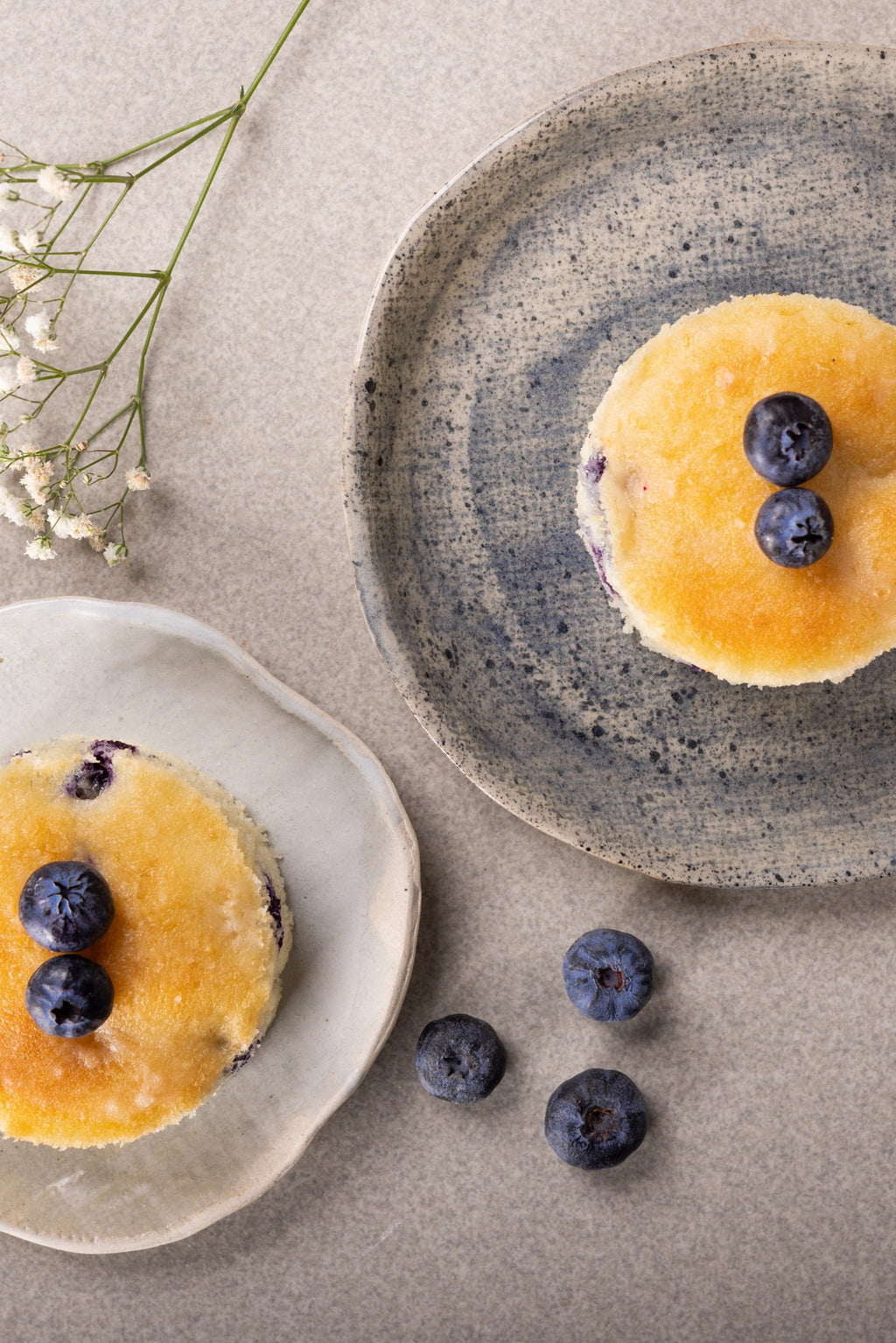 Two golden-brown blueberry tea cakes topped with fresh blueberries, presented on rustic ceramic plates on a neutral beige surface with small white flowers.