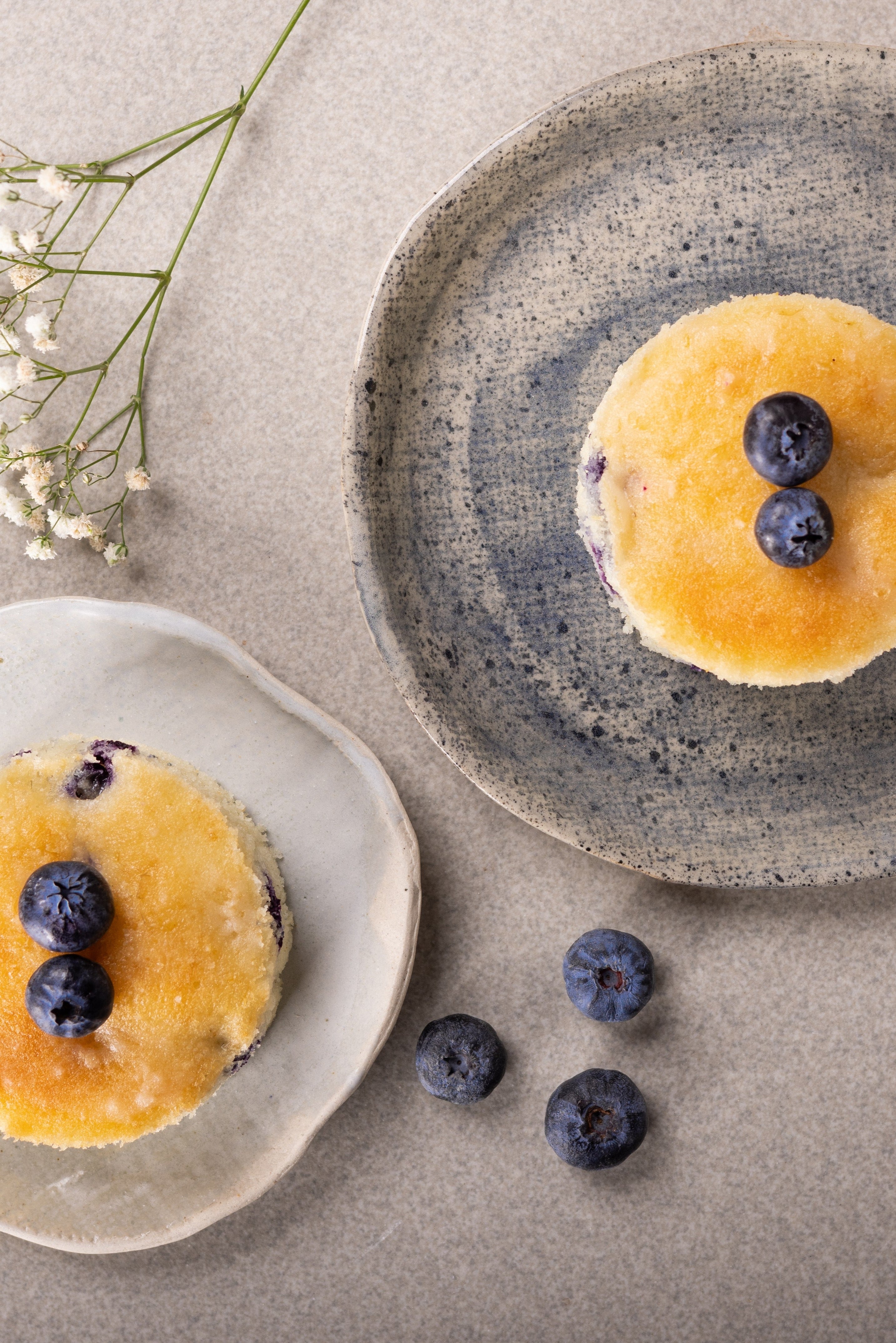 Two golden-brown blueberry tea cakes topped with fresh blueberries, presented on rustic ceramic plates on a neutral beige surface with small white flowers.