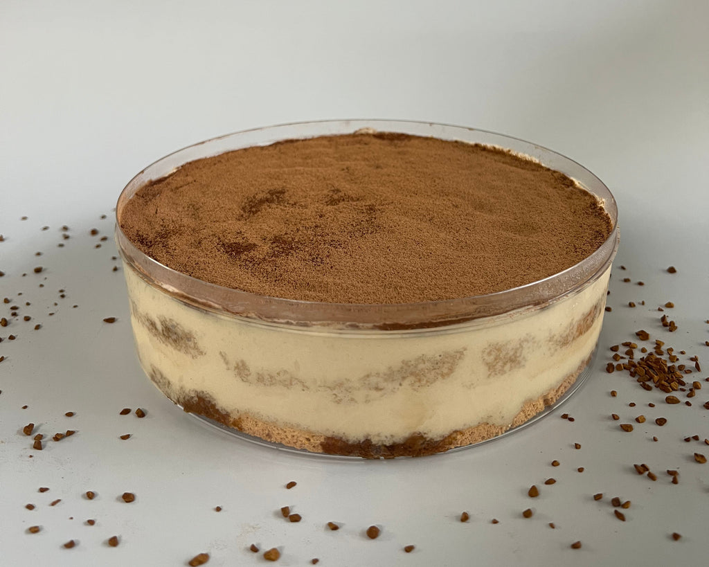 A round clear plastic tub filled with a layered tiramisu dessert, topped with cocoa powder, set on a light gray surface with cocoa crumbs scattered around.