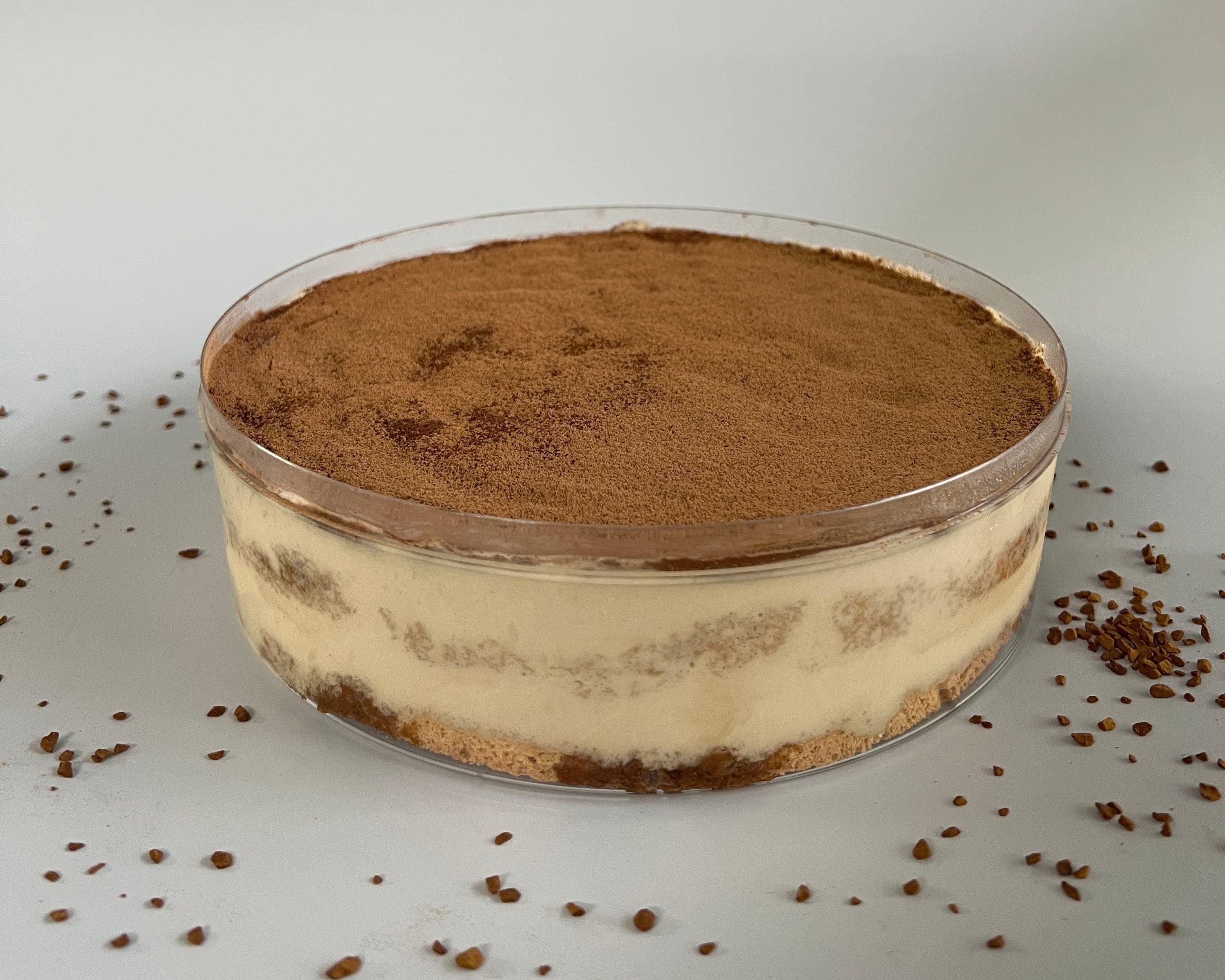 A round clear plastic tub filled with a layered tiramisu dessert, topped with cocoa powder, set on a light gray surface with cocoa crumbs scattered around.