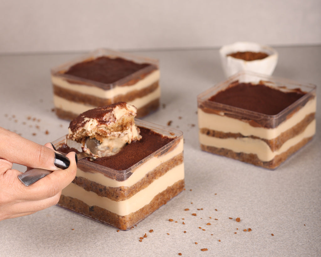 Three square-layered chocolate desserts with cream-colored layers in clear plastic containers, arranged on a light gray surface. A hand with black nail polish holds a spoonful of the dessert in the foreground, with two more desserts and a small white dish in the background.