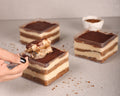Three square-layered chocolate desserts with cream-colored layers in clear plastic containers, arranged on a light gray surface. A hand with black nail polish holds a spoonful of the dessert in the foreground, with two more desserts and a small white dish in the background.